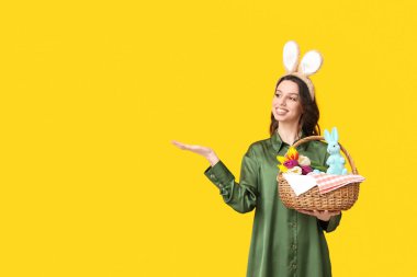 Paskalya tavşanı kulaklıklı mutlu genç kadın elinde laleler ve oyuncak tavşanla sarı arka plandaki bir şeyi gösteren sepet tutuyor.