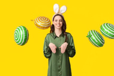 Paskalya tavşanı kulaklıklı, sarı arka planda balonları olan mutlu genç bir kadın.