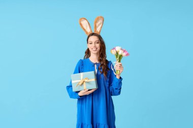 Paskalya tavşanı kulaklıklı, hediye kutusu ve mavi arka planda laleleri olan mutlu genç bir kadın.
