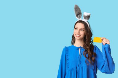 Paskalya tavşanı kulaklıklı, mavi arka planda kredi kartı olan mutlu genç bir kadın.