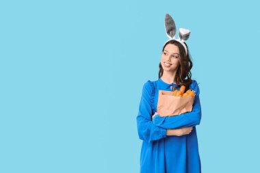 Paskalya tavşanı kulaklıklı mutlu genç kadın mavi arka planda havuçlu kağıt alışveriş torbası tutuyor.
