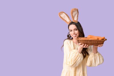 Paskalya tavşanı kulaklıklı mutlu genç kadın elinde havuçlu sepet ve eflatun arkaplan tutuyor.