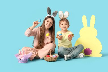 Erkek ve kız kardeşler oturmuş, ellerinde Paskalya yumurtası tutuyorlar mavi arka planda tavşanın yanında.