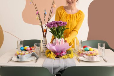 Paskalya sofrasını çiçek, mum ve boyalı yumurta dolu vazolarla hazırlayan kadın.