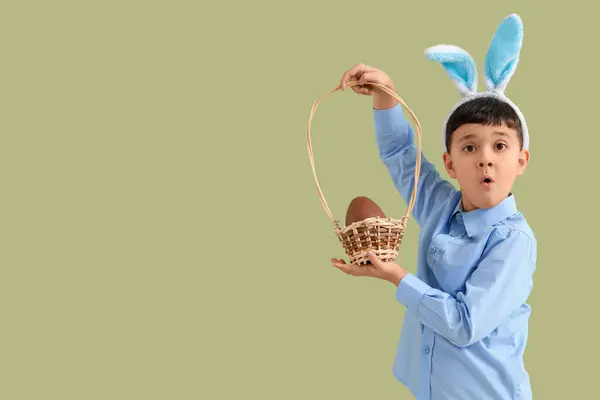緑の背景にイースターのためのチョコレートエッグとバスケットを保持するバニー耳の頭バンドで驚いた小さな少年 — ストック写真