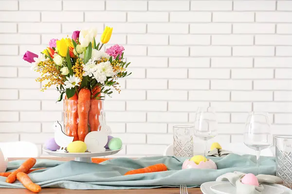 Beautiful table setting for Easter decorated with bouquet of carrots and flowers against white brick wall