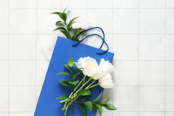 Paper shopping bag with flowers and green twigs on white tile background