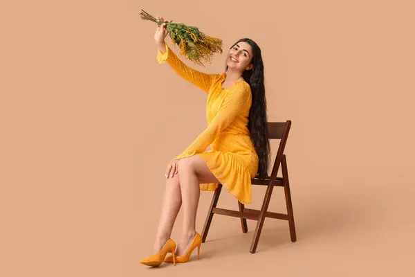 Beautiful young woman with bouquet of mimosa flowers sitting on chair against beige background