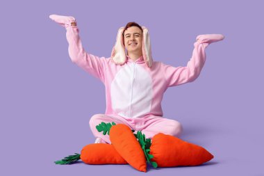 Paskalya tavşanı kostümlü mutlu genç adam peluş havuçlarla oturmuş leylak arka plandaki bir şeyi işaret ediyor.