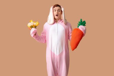 Paskalya tavşanı kostümlü genç adam elinde peluş havuç ve bej arka planda çiçekler tutuyor.