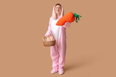 Paskalya tavşanı kostümlü mutlu genç adam elinde sepet ve bej arka planda peluş havuç tutuyor.