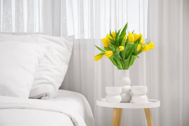 Yatak odasındaki komodinin üzerinde sarı laleler vazosu.