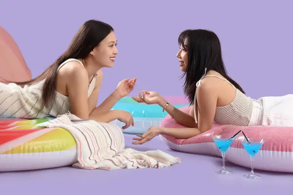 Female Asian friends chatting on swim mattresses against lilac background
