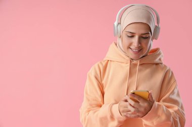 Pembe arka planda cep telefonu olan kulaklıklı genç Müslüman kadın.