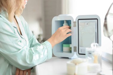 Kozmetik ürünleri için küçük bir buzdolabı olan güzel genç bir kadın yatak odasında oturuyor.