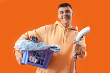 Buharlı tenceresi ve turuncu arka planda çamaşır dolu sepeti olan mutlu genç adam.