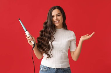Beautiful young woman with curling iron on red background