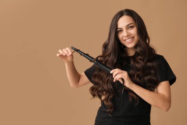 Beautiful young woman with curling iron on beige background