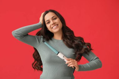 Beautiful young woman with curling iron on red background