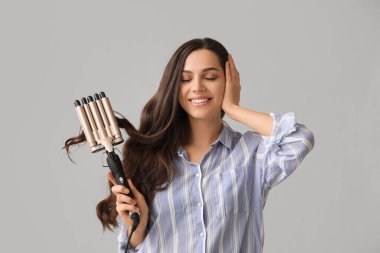 Beautiful young woman with curling iron on grey background