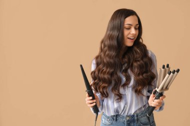 Beautiful young woman with different curling irons on beige background