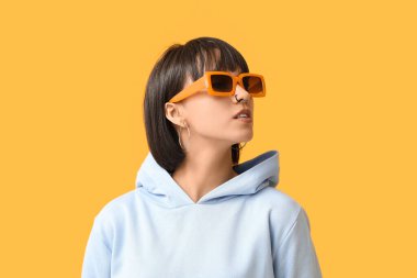 Sarı arka planda burun piercingli, güneş gözlüklü şık genç bir kadın.