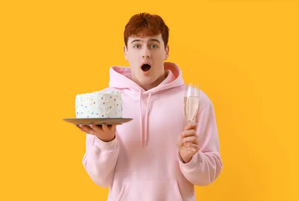 Shocked young man with birthday cake and glass of champagne on yellow background