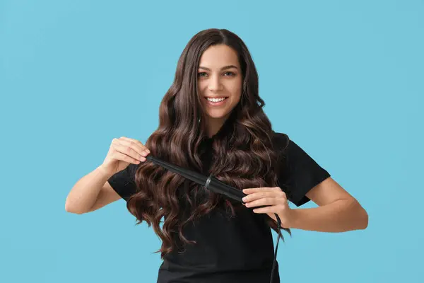 Beautiful young woman with curling iron on blue background