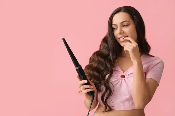 Beautiful young woman with curling iron on pink background