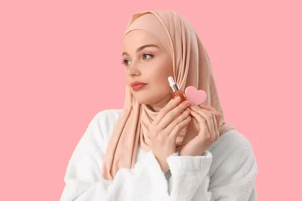 ピンクの背景にハート型のスポンジと血清を備えたヒジャブの美しいムスリム女性 クローズアップ — ストック写真