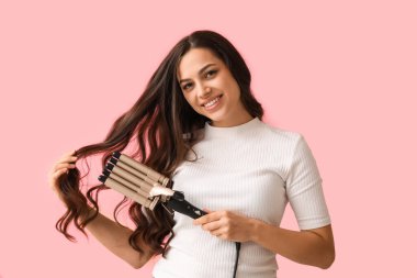 Beautiful young woman with curling iron on pink background