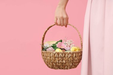 Elinde makyaj malzemeleri, çiçekler ve pembe arka planda Paskalya dekoru olan sepet tutan genç bir kadın.