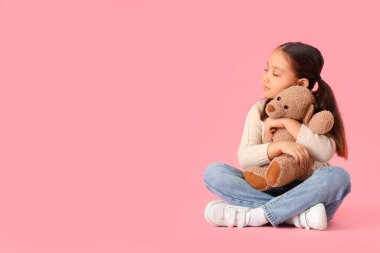 Otistik bozukluğu olan küçük Asyalı kız pembe arka planda oyuncak ayıya sarılıyor.