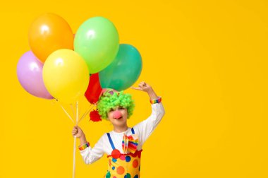 Sarı arka plandaki balonları gösteren palyaço kostümlü komik çocuk. 1 Nisan şakası