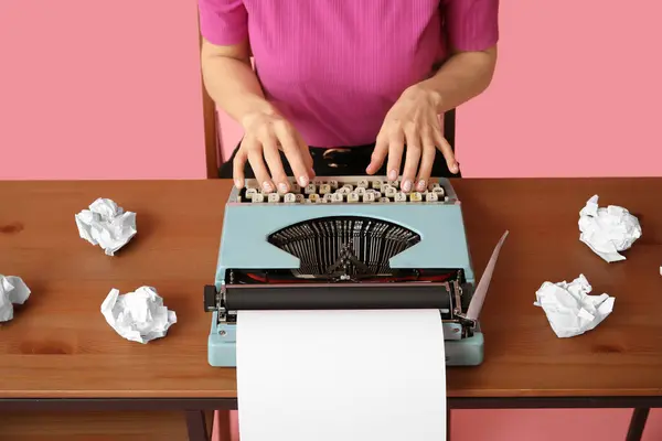 Young woman with vintage typewriter at table on pink background