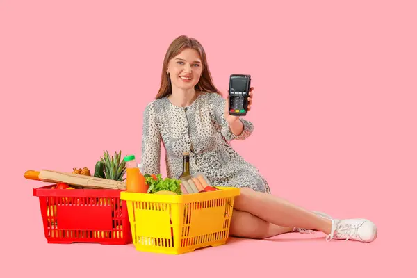 Young woman with payment terminal and full shopping baskets on pink background