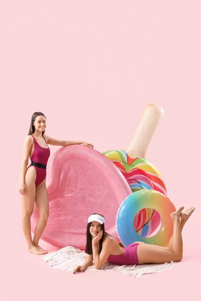 Female friends in beachwear with swim mattresses on pink background