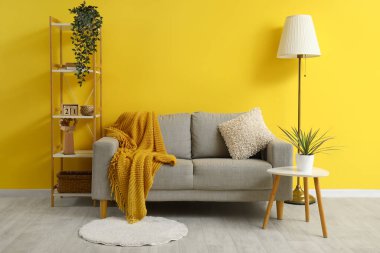 Ekose ve yastık gri kanepede, lamba, masa ve ahşap askı sarı duvarın yanında