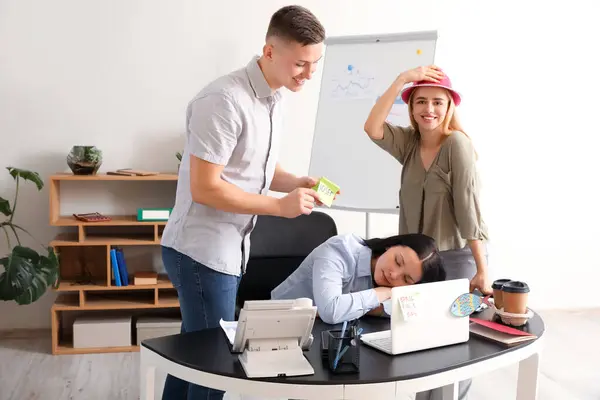 Business people sticking paper on their sleeping colleague in office. April Fools\' Day celebration