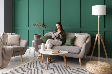 Güzel bir kadın oturma odasındaki gri koltukta dergi okuyor.