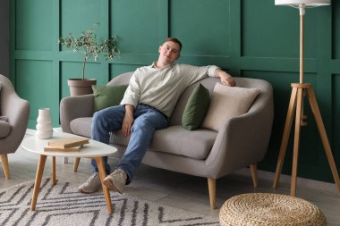 Genç adam oturma odasındaki gri kanepede dinleniyor.