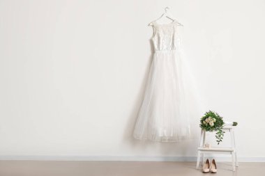 Beyaz duvarda asılı güzel bir gelinlik, gelinlik ayakkabıları ve gelin buketi.