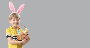 Tavşan kulaklı şirin bir çocuk elinde sepetle gri arka planda oyuncak tavşanlar ve mesaj için yeri var. Paskalya kutlaması