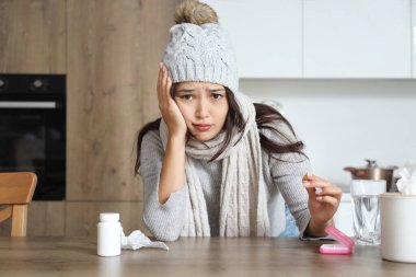 Sick Asian woman with pills and tissues sitting at table in kitchen clipart