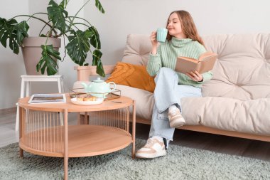 Kitaplı genç kadın evdeki kanepede çay içiyor.