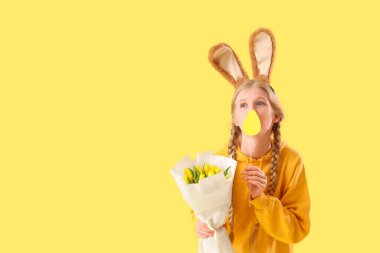 Paskalya tavşanı kulakları, çiçekleri ve sarı arka planda kağıt yumurtası olan genç bir kız.