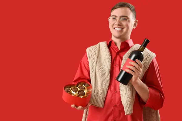 Young man with heart-shaped chocolate candies and wine on red background. Valentine\'s Day celebration