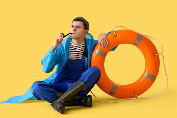 Young sailor in raincoat with smoking pipe and ring buoy on yellow background