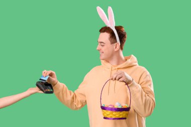 Tavşan kulaklı, sepetli genç adam yeşil arka planda terminalden ödeme yapıyor. Paskalya Satışı