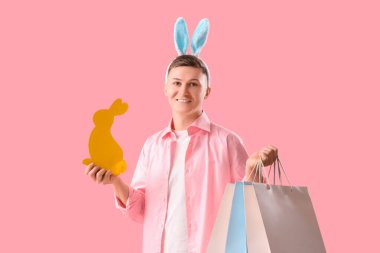 Alışveriş torbaları ve pembe arka planda kağıt tavşanlı tavşan kulaklı genç adam. Paskalya Satışı